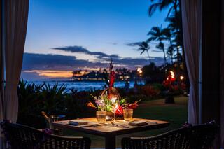 A cozy dining setup by the ocean at sunset, adorned with vibrant flowers and soft lighting, creates a romantic and tranquil atmosphere.