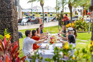 A cheerful group toasts with drinks at an outdoor table surrounded by lush greenery and a pool, enjoying a festive gathering.