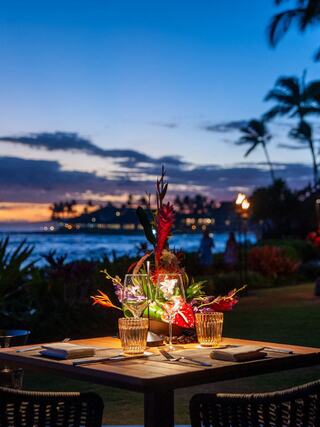 A romantic outdoor dining setup with tropical flowers, illuminated glasses, and a serene sunset over the ocean in the background.