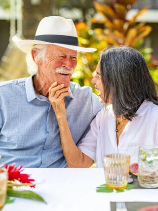A joyful couple shares a moment of laughter and affection in a vibrant outdoor setting, surrounded by tropical plants and a festive dining table.