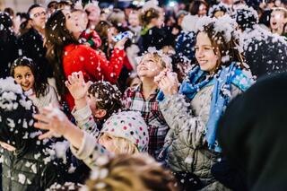 Joyful children and adults celebrate in a festive atmosphere, surrounded by falling artificial snow, creating a magical winter experience.