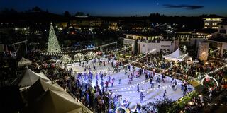 A vibrant outdoor ice skating rink is bustling with skaters, surrounded by festive lights and a large Christmas tree under a twilight sky.