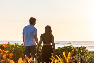 A couple stands hand-in-hand, gazing at the ocean under a warm sunset, surrounded by vibrant greenery.