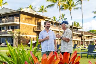 Two men enjoy a friendly conversation in a tropical setting, surrounded by vibrant flowers and palm trees, with a resort backdrop.