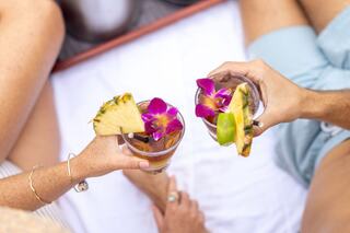 Two hands hold tropical drinks adorned with flowers and fruit, set against a light background, suggesting a relaxing moment together.