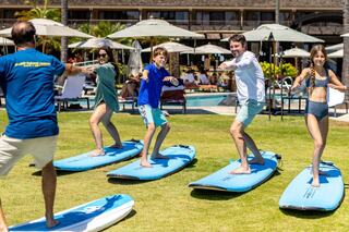 A family enjoys a surfing lesson on land, balancing on surfboards while a coach guides them under sunny skies at a resort.