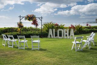 A serene beachside setting features white chairs arranged in a circle, lush greenery, and a "ALOHA" sign, perfect for a celebration.