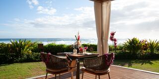 A cozy outdoor dining setup overlooks a beautiful ocean, with colorful flowers surrounding the table and a bright sky above.