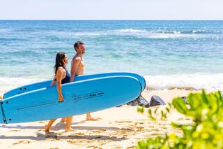 A man and woman carry surfboards along a sandy beach, with sunny skies and gentle waves in the background, suggesting a fun day of surfing ahead.