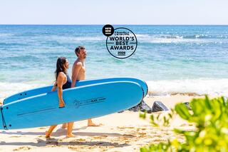 Two people carry bright blue surfboards along a sandy beach, enjoying a sunny day with the ocean waves in the background.