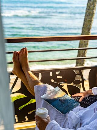 Bare feet relax on a balcony overlooking the ocean, with a coffee cup in one hand and a magazine in the other, embodying tranquility.