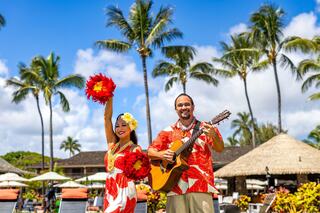 A woman twirls with flowers, while a man strums a guitar, set against a tropical paradise with palm trees and a resort backdrop.