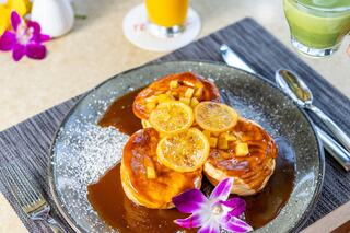 Delicious pancakes topped with bananas and drizzled with syrup, garnished with flowers, alongside vibrant drinks. A tropical breakfast delight!