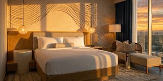 A stylish hotel room features a king bed, cozy seating, and large windows with a stunning sunset view, exuding warmth and modern comfort.