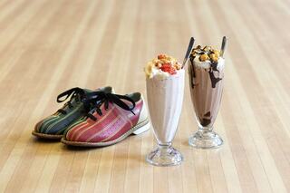 Colorful bowling shoes sit next to two delicious milkshakes topped with whipped cream and garnishes, on a wooden floor.