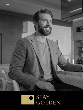A smiling man in a stylish blazer sits at a bar, exuding confidence and charm in a modern, elegant setting.