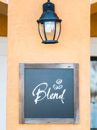 A warm orange wall features a stylish sign that reads "Blend," adorned with coffee bean illustrations and complemented by a lantern above.