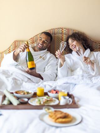 A couple in plush robes enjoys champagne and breakfast in bed, sharing laughter and a cozy, intimate moment.