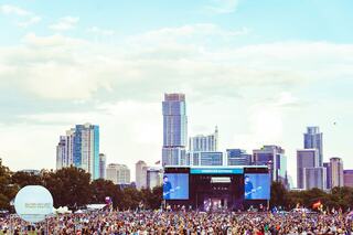 A vibrant crowd gathers in an open space, with a large stage set against a skyline of modern buildings under a cloudy sky.