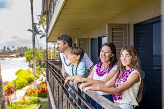 A family enjoys a sunny day on a balcony, adorned with colorful leis, smiling and admiring a scenic view of lush greenery and ocean.