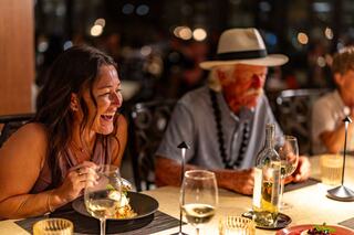 A woman laughs heartily over dinner, while an older man in a hat enjoys the moment. Soft lighting and elegant table settings create a warm atmosphere.