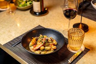 A gourmet dish of seafood with vibrant garnishes sits in a stylish bowl, accompanied by a glass of red wine and a golden tumbler.