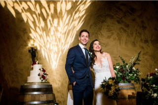 A smiling couple poses joyfully in a romantic venue, surrounded by elegant decor, a wedding cake, and beautiful floral arrangements.