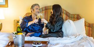 A couple in stylish pajamas toasts with champagne in a cozy, beautifully arranged hotel room, enjoying a romantic moment together.