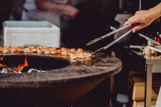 A chef uses tongs to grill skewered meat on a circular cooking surface, with flames visible and a blurred kitchen background.