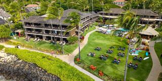 A tropical resort features lush green lawns, palm trees, a pool area, and cozy lounges, with buildings nestled amidst vibrant flora.