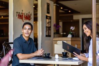 A man and woman sit at a café table, smiling and sharing a moment, with menus and coffee cups in a stylish, relaxed atmosphere.