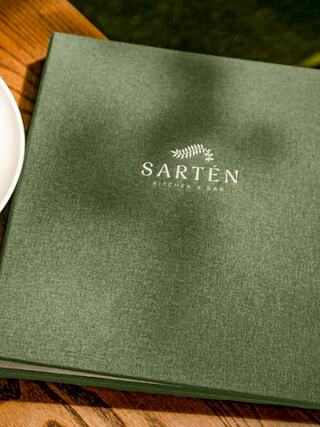A green menu for SARTÉN Kitchen & Bar rests on a wooden table, accompanied by a white plate, suggesting a cozy dining atmosphere.