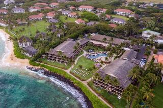 Aerial view of a tropical resort by the beach, featuring lush greenery, swimming pools, and beachfront access surrounded by palm trees.