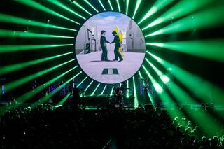 A vibrant concert scene features a large circular screen displaying animated figures, surrounded by beams of green light and an engaged audience.