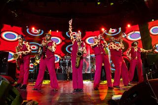 A lively band in vibrant red outfits performs on stage, surrounded by colorful retro patterns and energetic lighting.