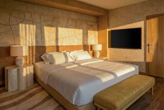 A cozy bedroom features a large bed, stylish lamps, a flat-screen TV, and warm sunlight creating a serene atmosphere.