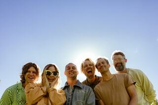 A group of six smiling friends stands together outdoors, illuminated by sunlight with a clear blue sky behind them, radiating joy and camaraderie.