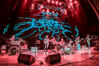 A lively band performs on stage under vibrant lights, with dynamic visuals projected behind them, creating an energetic atmosphere.