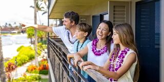 A family in Hawaiian leis enjoys a scenic view from their balcony, smiling and sharing a joyful moment together.