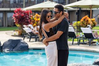A couple embraces by a pool, enjoying a sunny day, surrounded by vibrant tropical plants and lounge chairs in the background.