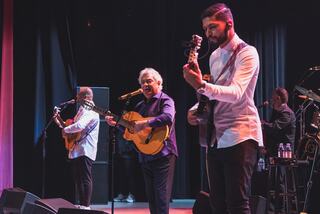 Three musicians perform on stage, playing guitars and singing, with colorful lighting and an enthusiastic atmosphere.