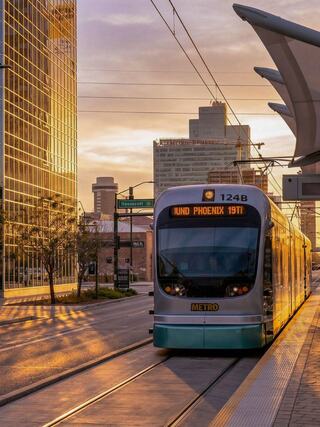 A light rail tram approaches a station at sunrise, with modern buildings reflecting golden light in a vibrant urban setting.