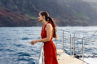 A woman in a red dress leans against a boat railing, gazing thoughtfully at the ocean and rocky cliffs in the background.