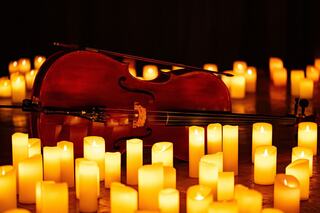 A cello lies surrounded by glowing candles, creating an intimate and serene atmosphere. Warm light enhances the wooden tones of the instrument.