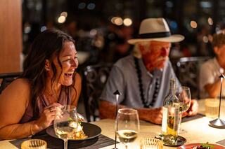 A woman laughs joyfully while dining, sharing a table with an older man in a hat and people enjoying a vibrant evening atmosphere.
