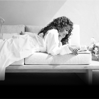A woman in a cozy robe lies on a couch, reading contently, surrounded by flowers and soft light illuminating her peaceful moment.