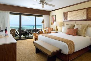A coastal hotel room features a king bed with decorative pillows, a comfortable chair, and a balcony overlooking the ocean. Natural light pours in.