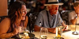 A woman joyfully enjoys her meal at a dinner table, while an older man in a hat sits nearby, creating a lively dining atmosphere.
