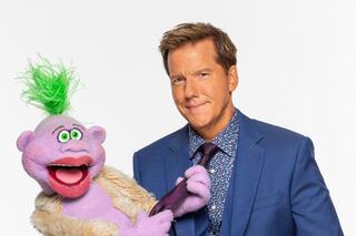 A man in a suit poses with a colorful puppet featuring green hair and expressive eyes, both looking playfully at the camera.