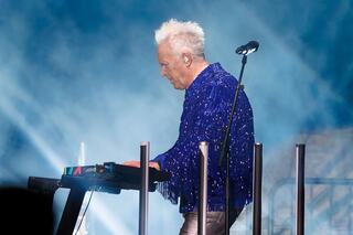 A musician in a sparkly blue jacket performs on stage, focused on their synthesizer amid a misty, illuminated atmosphere.